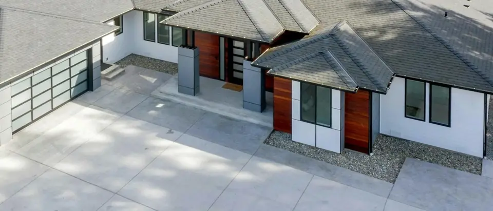 A single-story house with gray asphalt shingle hip roofs, white walls with wood panels and a wide concrete driveway, seen from above