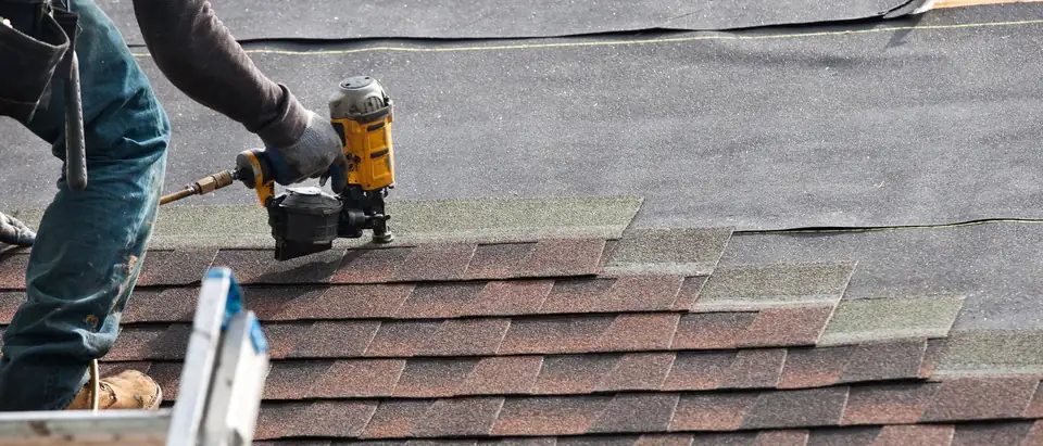 A roofer's gloved hand driving nails into red-brown asphalt shingles with a coil nail gun, with roofing felt on the deck above