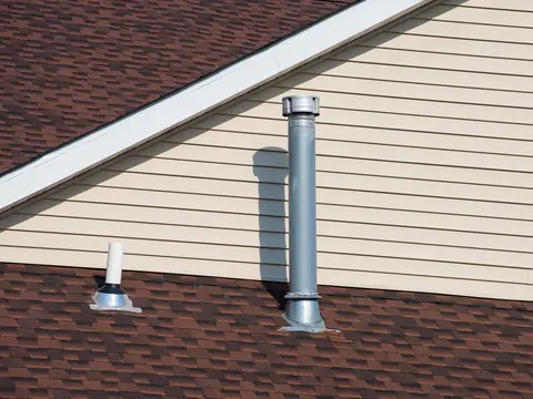 A white plastic vent pipe with a rubber collar and a metal flashing base, and a taller metal flue beside it, on a red-brown shingle roof