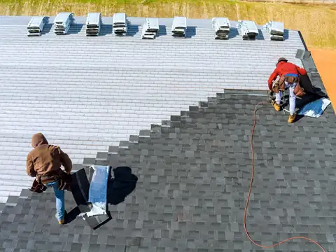 Two roofers seen from above laying gray asphalt shingles in stepped rows over white underlayment
