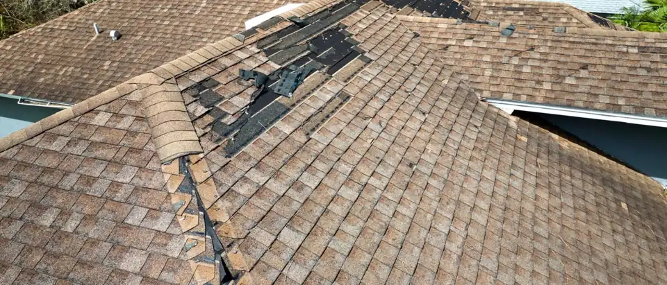 A wind-damaged brown asphalt shingle roof with ridge caps peeled back along a hip and a patch of shingles torn off